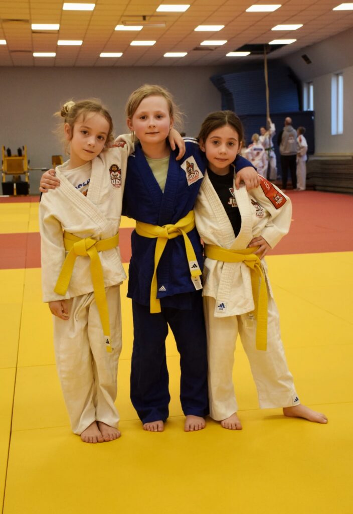 Trzy dziewczynki w białych i niebieskim judogu Judo Lion z żółtymi pasami stoją objęte na żółtych matach w sali.