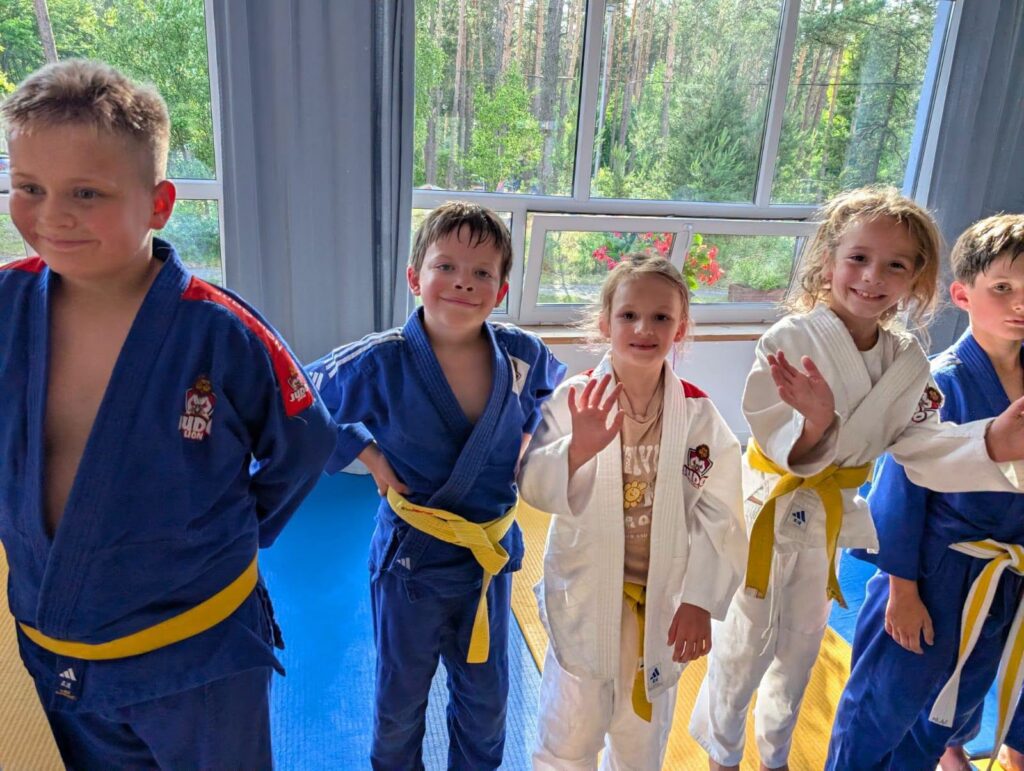 Pięcioro dzieci w niebieskich i białych judogach Judo Lion z żółtymi pasami uśmiecha się i macha do aparatu przy dużych oknach.