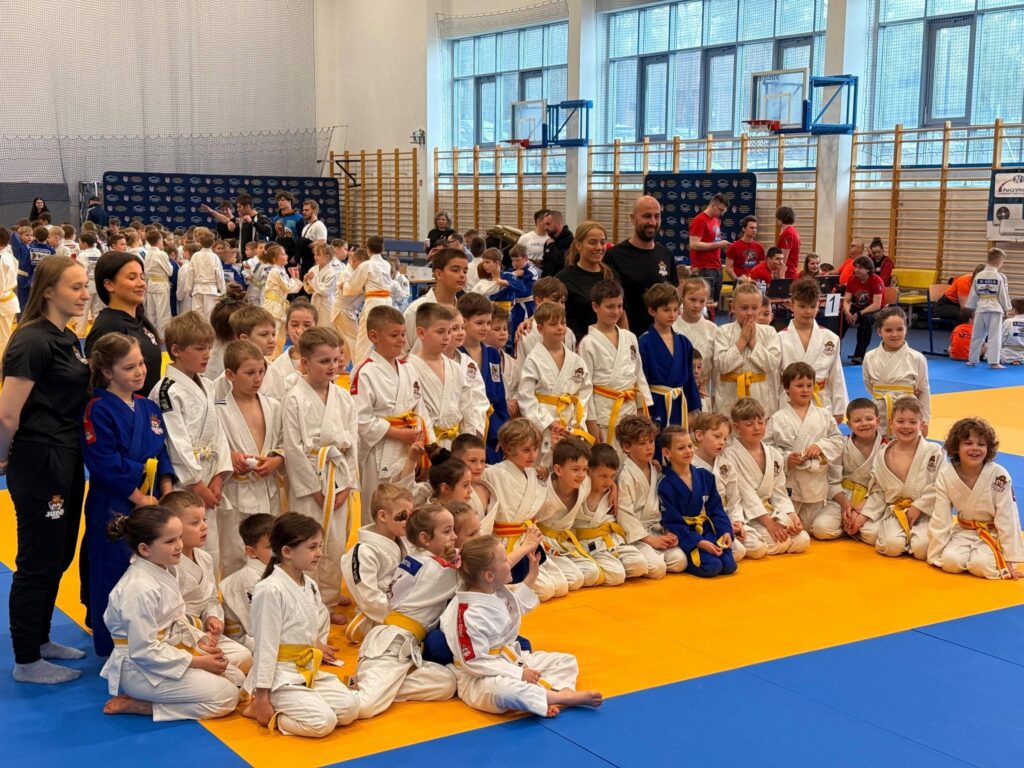 Liczna grupa dzieci w białych i niebieskich judogach z trenerkami pozuje na żółto-niebieskich matach w dużej hali sportowej.